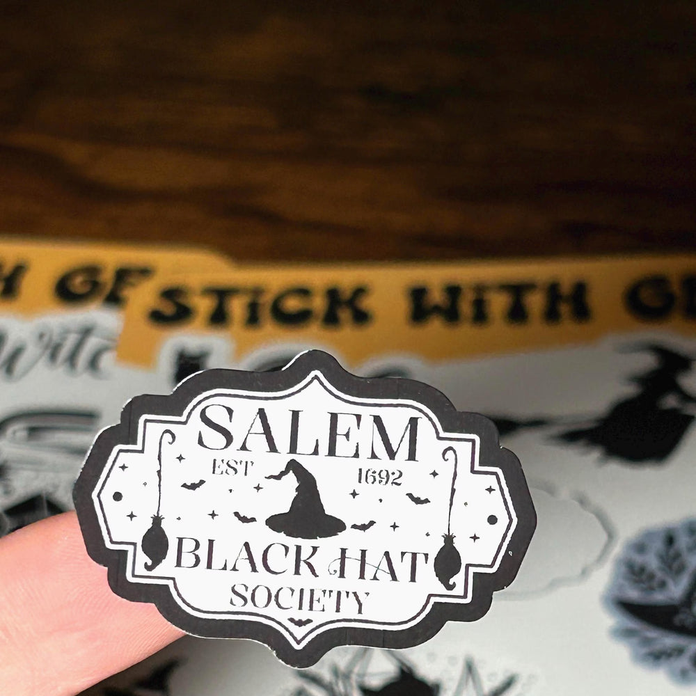 Sticker with 'Salem Black Hat Society' design held by a hand, with a sticker sheet in the background.