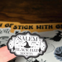 Load image into Gallery viewer, Sticker with 'Salem Black Hat Society' design held by a hand, with a sticker sheet in the background.
