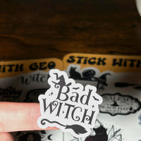Load image into Gallery viewer, Sticker with 'Bad Witch' text held over a sheet of stickers with similar designs.
