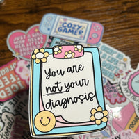 Load image into Gallery viewer, Sticker with 'You are not your diagnosis' text on a wooden surface with other stickers.
