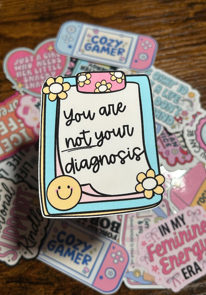 Sticker with 'You are not your diagnosis' text on a wooden surface with other stickers.