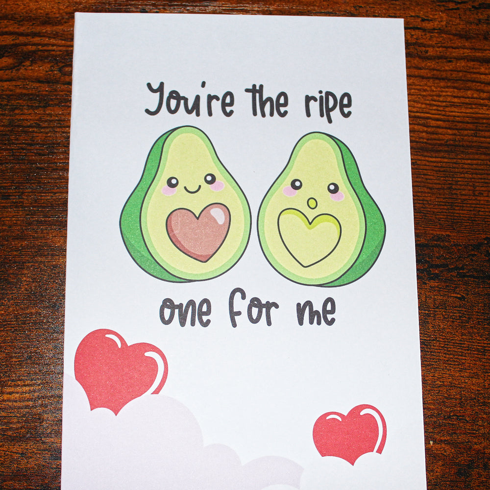 Card with cartoon avocados and text 'You're the ripe one for me' on a wooden surface