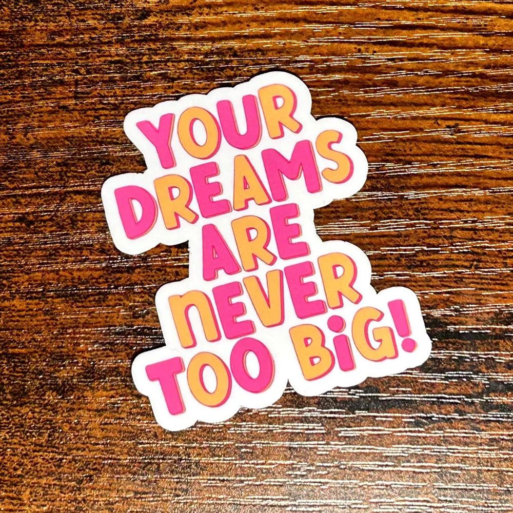 Sticker with motivational text 'Your dreams are never too big!' on a wooden surface