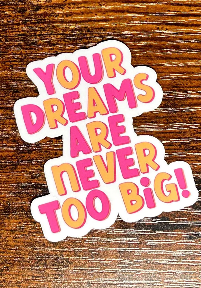 Sticker with motivational text 'Your dreams are never too big!' on a wooden surface