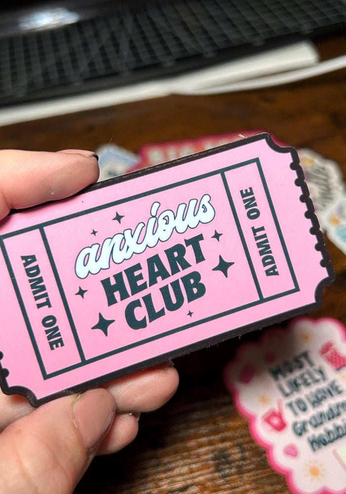 Pink ticket-shaped sticker with 'anxious HEART CLUB' text held by a hand on a wooden surface.