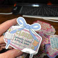 Load image into Gallery viewer, Sticker with a blue bow and text held by a hand on a wooden surface with other stickers in the background.
