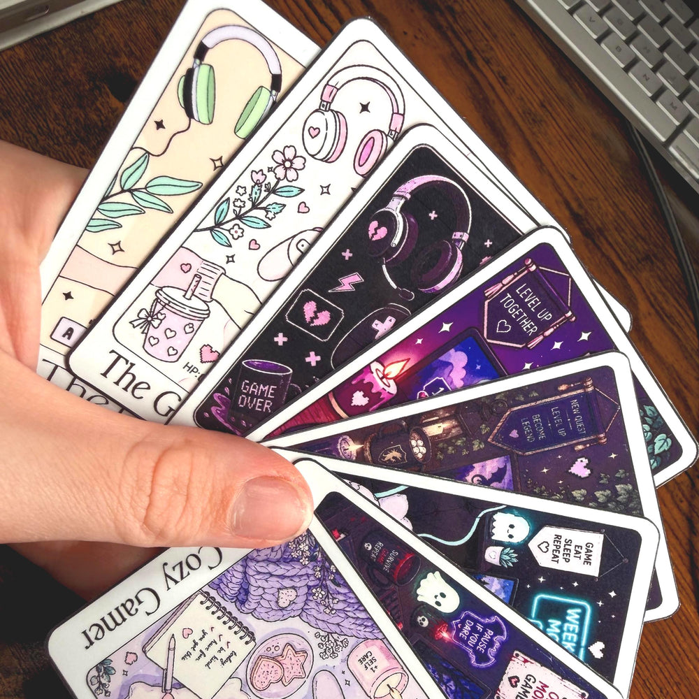 A hand holds a fan of illustrated stickers with gaming and cozy aesthetic designs, photographed on a wooden desk near a keyboard.