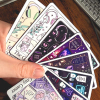 Load image into Gallery viewer, A hand holds a fan of illustrated stickers with gaming and cozy aesthetic designs, photographed on a wooden desk near a keyboard.
