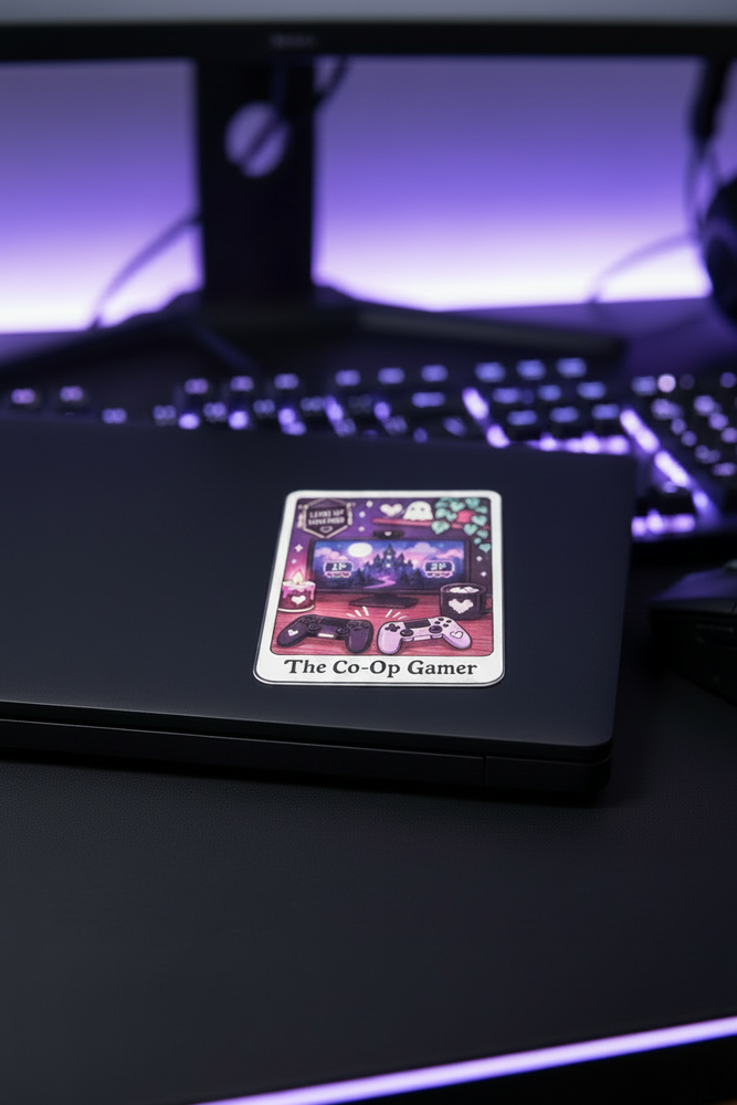 A square sticker with a colorful cartoon illustration and the text 'The Co-Op Gamer' is shown on the lid of a black laptop. The laptop sits on a desk in front of a blurred computer keyboard and monitor with purple backlighting.