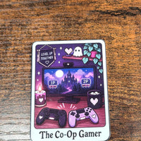 Load image into Gallery viewer, A sticker featuring a cute pixel-art illustration titled 'The Co-Op Gamer' resting on a dark wood surface. The illustration depicts a spooky gaming scene with a TV screen, two game controllers, candles, and ghosts.
