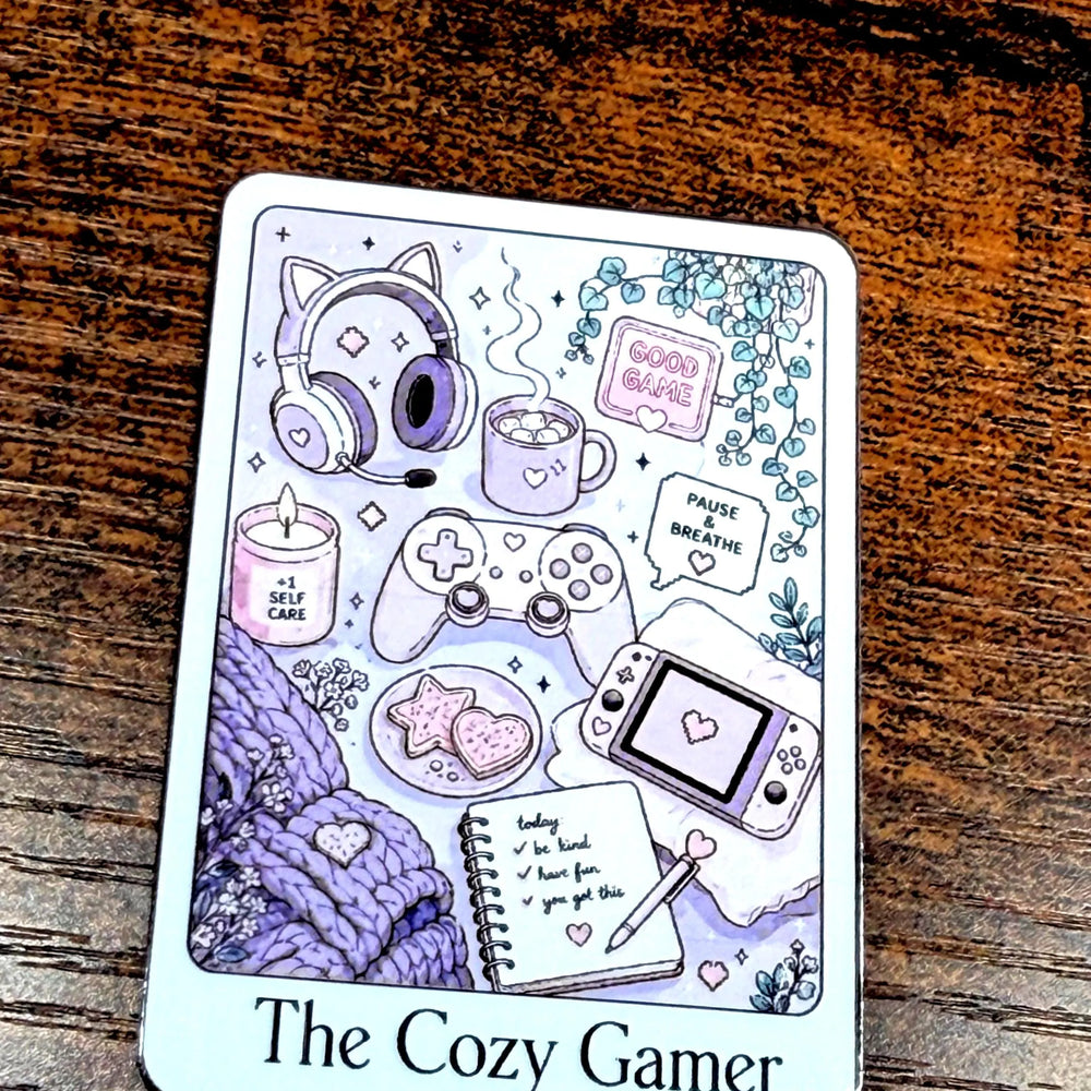 A rectangular illustrated Sticker labeled 'The Cozy Gamer' sits on a dark wooden surface. The front shows pastel drawings of gaming-themed items including headphones, a game controller, a handheld console, a candle, a notebook, and cozy textures in lavender, purple, pink, and white.