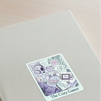 Load image into Gallery viewer, A beige hardcover book or notebook lying on a light wood tabletop, with a decorative sticker on the cover that reads 'The Cozy Gamer' and features pastel gaming-themed illustrations.
