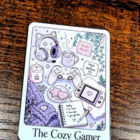 Load image into Gallery viewer, A rectangular illustrated Sticker labeled 'The Cozy Gamer' sits on a dark wooden surface. The front shows pastel drawings of gaming-themed items including headphones, a game controller, a handheld console, a candle, a notebook, and cozy textures in lavender, purple, pink, and white.
