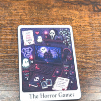 Load image into Gallery viewer, A tarot sticker titled 'The Horror Gamer' featuring a dark, cozy gaming scene with ghosts, a laptop, headphones, and a haunted house, displayed on a wooden tabletop.
