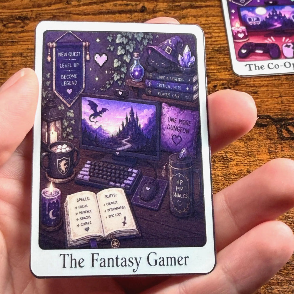 A hand holds a tarot-style Sticker titled 'The Fantasy Gamer' featuring a purple and black illustration of a gaming setup in a fantasy setting. Another Sticker is partially visible on the wooden table in the background.