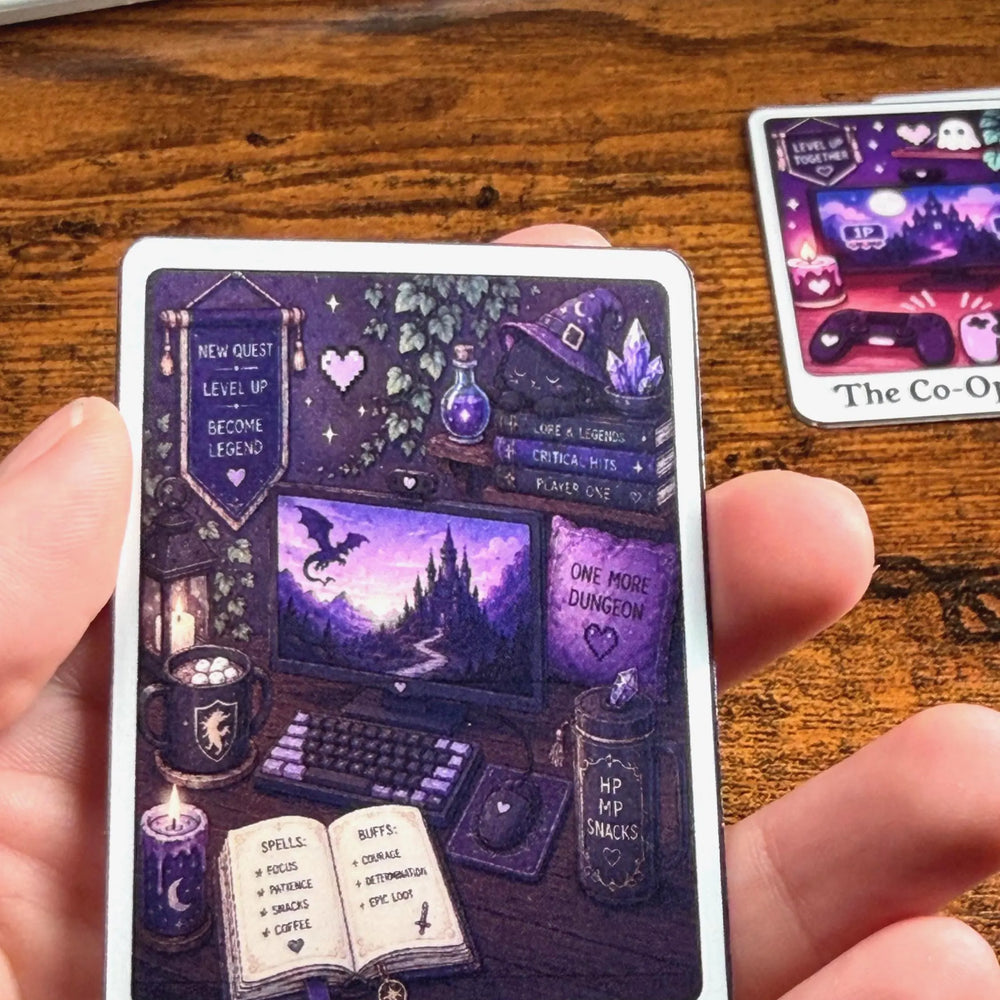 A hand holds a tarot-style Sticker titled 'The Fantasy Gamer' featuring a purple and black illustration of a gaming setup in a fantasy setting. Another Sticker is partially visible on the wooden table in the background.