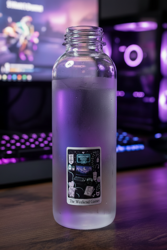 A clear glass water bottle with a white sticker, filled with water and sitting on a wooden desk with a blurred gaming setup in the background.
