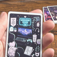 Load image into Gallery viewer, A hand holds a rectangular printed Sticker titled "The Weekend Gamer" against a wooden tabletop, with two more stickers partially visible in the background.
