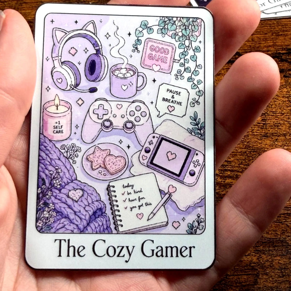 A hand holding a rectangular Sticker  titled 'The Cozy Gamer' featuring cute purple and lavender gaming-themed illustrations, including a controller, headset, mug, game console, notebook, and cozy accessories, set against a wooden tabletop background.