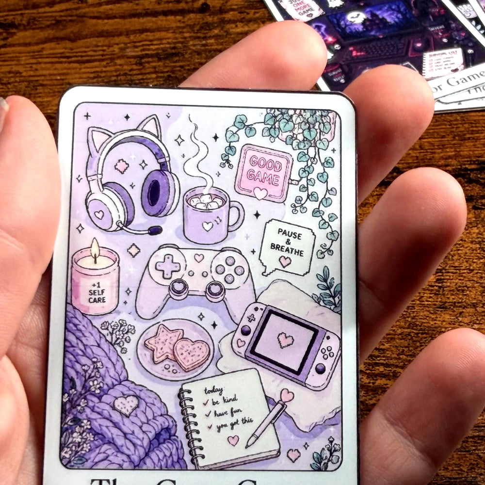 A hand holding a rectangular Sticker  titled 'The Cozy Gamer' featuring cute purple and lavender gaming-themed illustrations, including a controller, headset, mug, game console, notebook, and cozy accessories, set against a wooden tabletop background.