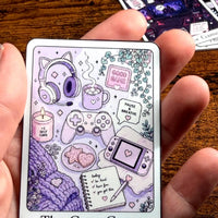 Load image into Gallery viewer, A hand holding a rectangular Sticker  titled 'The Cozy Gamer' featuring cute purple and lavender gaming-themed illustrations, including a controller, headset, mug, game console, notebook, and cozy accessories, set against a wooden tabletop background.
