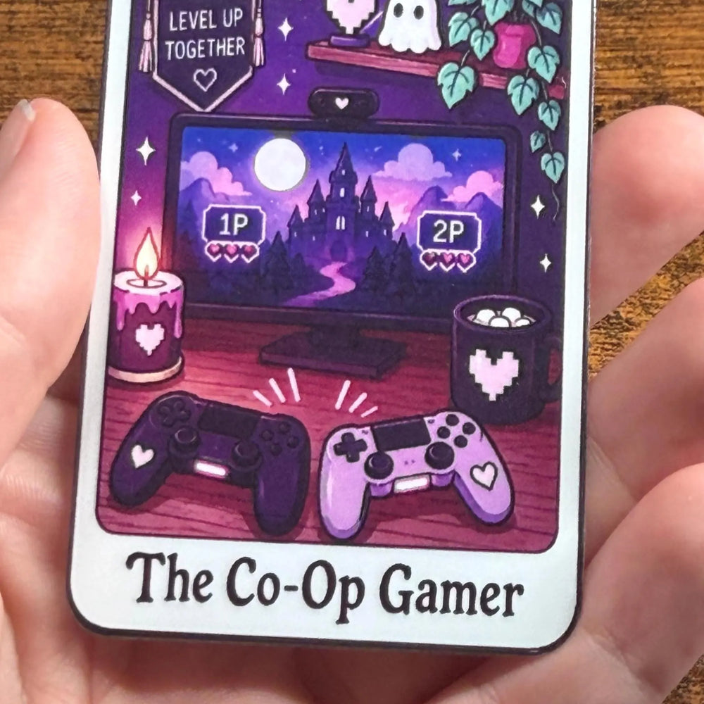 A hand holding a rectangular sticker featuring a purple-themed gaming illustration with the text 'The Co-Op Gamer'. The sticker shows a computer monitor with a castle in the distance, two game controllers, candles, a mug, and ghosts, all in shades of purple, pink, and black.