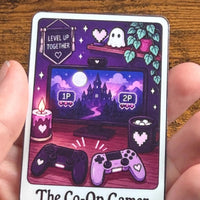 Load image into Gallery viewer, A hand holding a rectangular sticker featuring a purple-themed gaming illustration with the text 'The Co-Op Gamer'. The sticker shows a computer monitor with a castle in the distance, two game controllers, candles, a mug, and ghosts, all in shades of purple, pink, and black.
