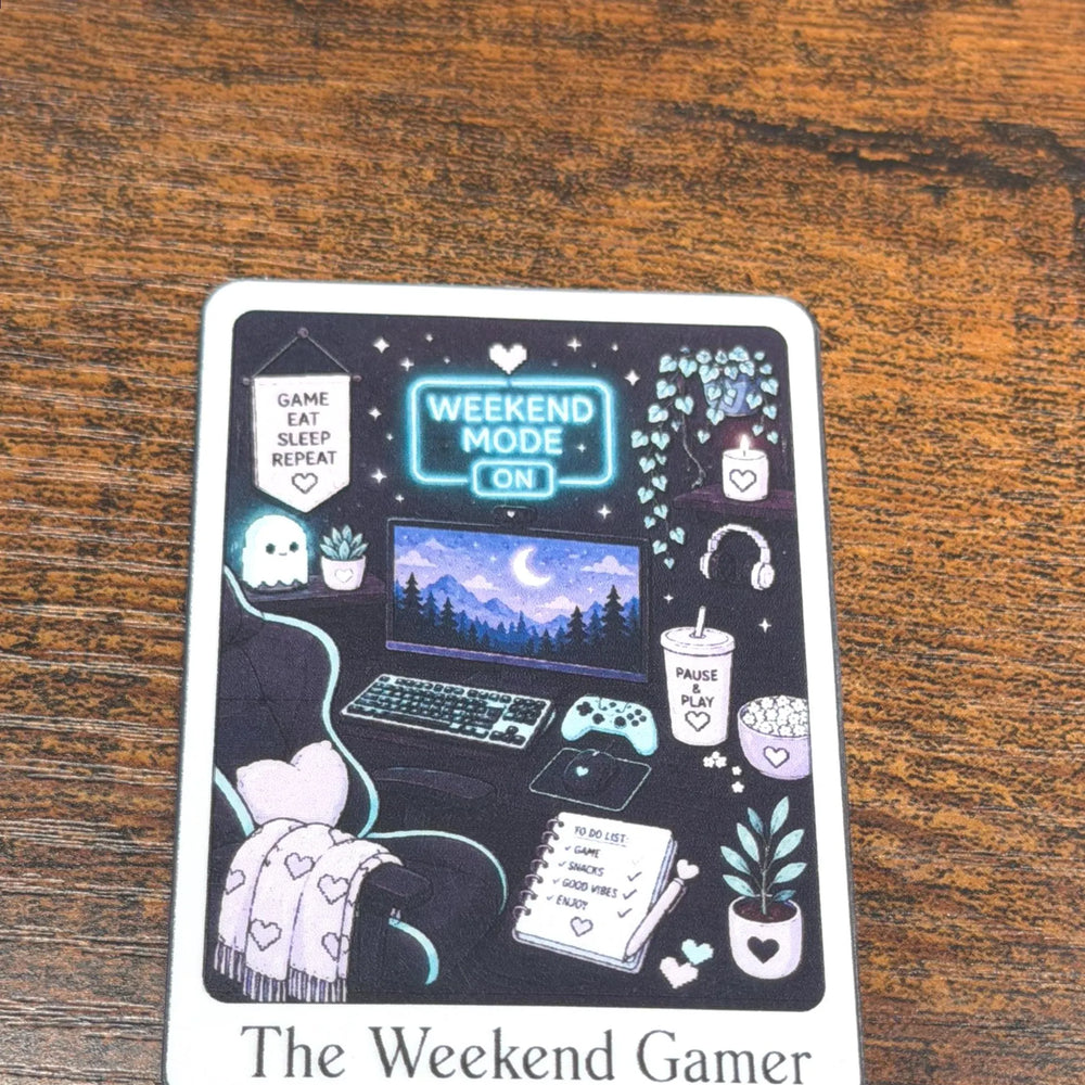 A square printed sticker with rounded corners sits on a wooden tabletop. The design features a dark illustrated scene labeled 'The Weekend Gamer' with a laptop, gaming setup, plants, headphones, and cozy decor.