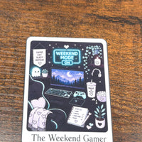 Load image into Gallery viewer, A square printed sticker with rounded corners sits on a wooden tabletop. The design features a dark illustrated scene labeled 'The Weekend Gamer' with a laptop, gaming setup, plants, headphones, and cozy decor.
