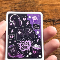 Load image into Gallery viewer, A hand holding a rectangular printed sticker titled "The Rage Quitter," featuring a dark purple gaming-themed illustration with headphones, controllers, hearts, and text like "RAGE QUIT" and "GAME OVER," against a wooden tabletop background.
