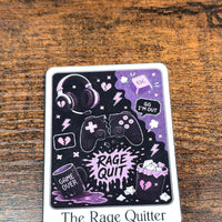 Load image into Gallery viewer, A rectangular printed  sticker titled 'The Rage Quitter' with a black and purple gaming-themed illustration, placed on a brown wooden surface.
