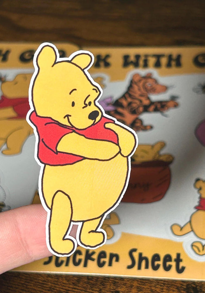 Sticker of a yellow bear with a red shirt held over a Winnie the Pooh sticker sheet.
