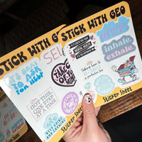 Load image into Gallery viewer, Sticker sheet with various designs and text held by a hand on a wooden surface.
