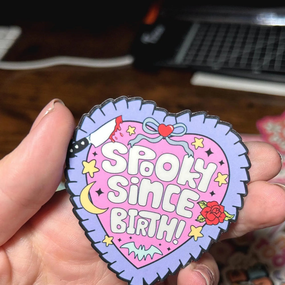 Heart-shaped patch with 'Spook Since Birth' text held by a hand on a wooden surface.