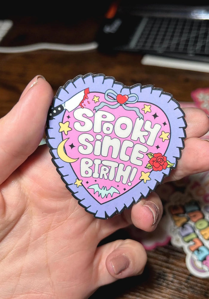 Heart-shaped patch with 'Spook Since Birth' text held by a hand on a wooden surface.